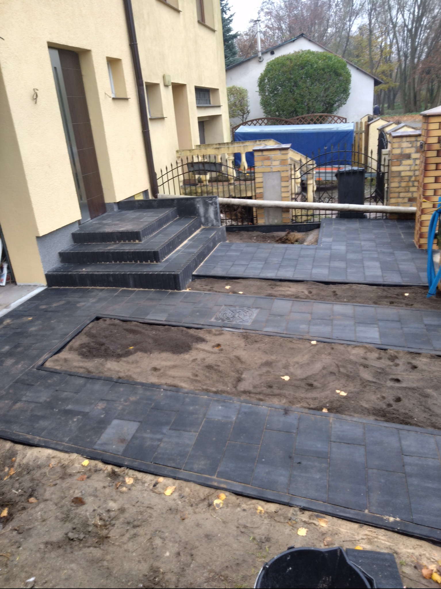 Ułożona czarna kostka brukowa przed wejściem do domu z żółtą elewacją, widoczne schody i fragmenty rabat ziemnych, w tle ogrodzenie i zabudowania.