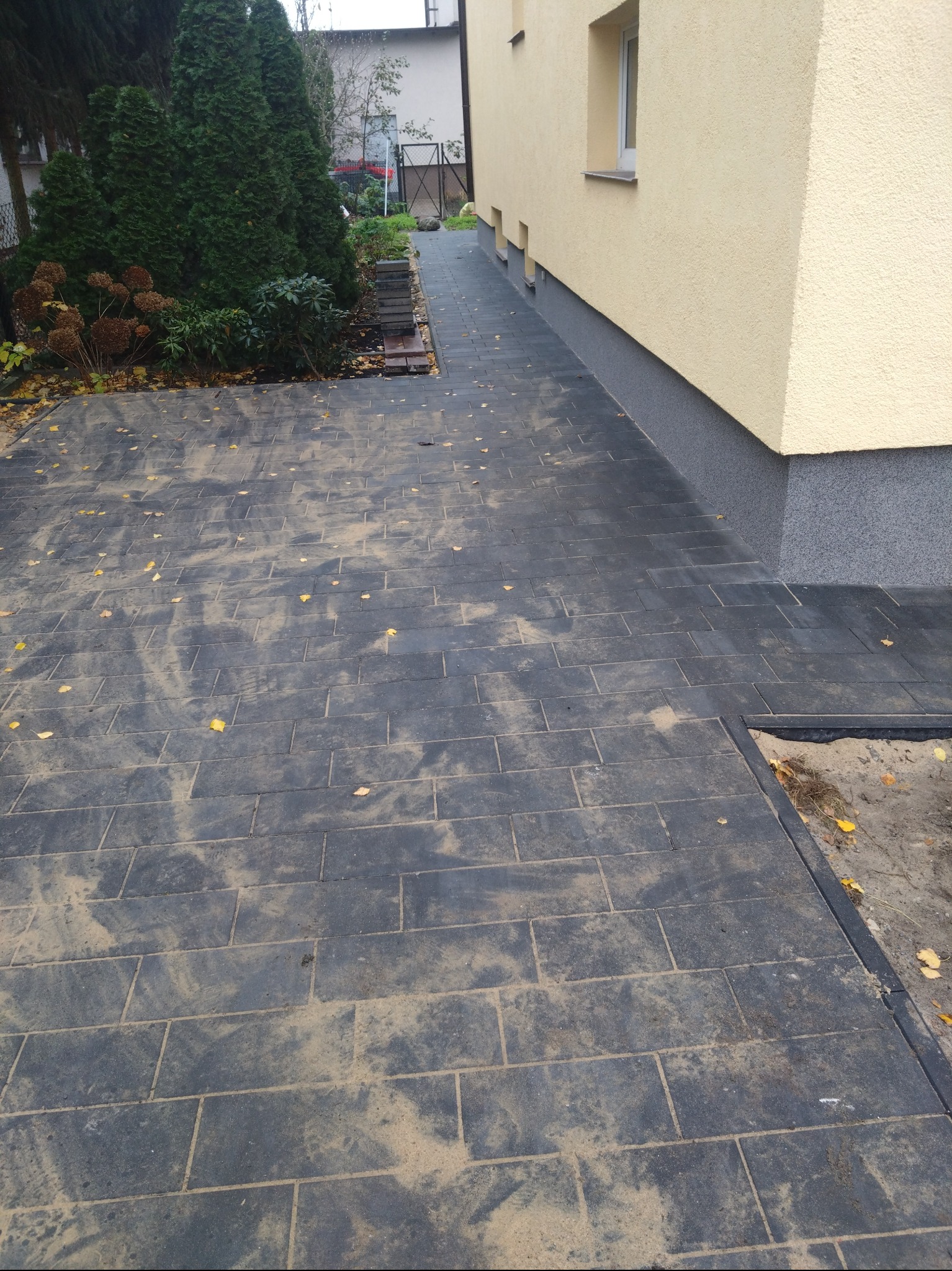 Ułożona, grafitowa kostka brukowa z fugami wypełnionymi piaskiem, prowadząca wzdłuż ocieplonej elewacji budynku w kolorze beżowym z szarym cokołem, z widocznymi drzewami iglastymi w tle.