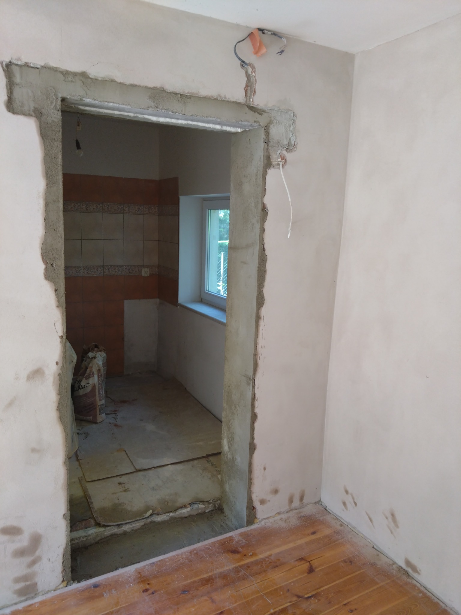 Przygotowanie do montażu ościeżnicy: surowy beton wokół otworu drzwiowego, widoczne kable elektryczne, fragment wykończonej łazienki w tle i worki z zaprawą.