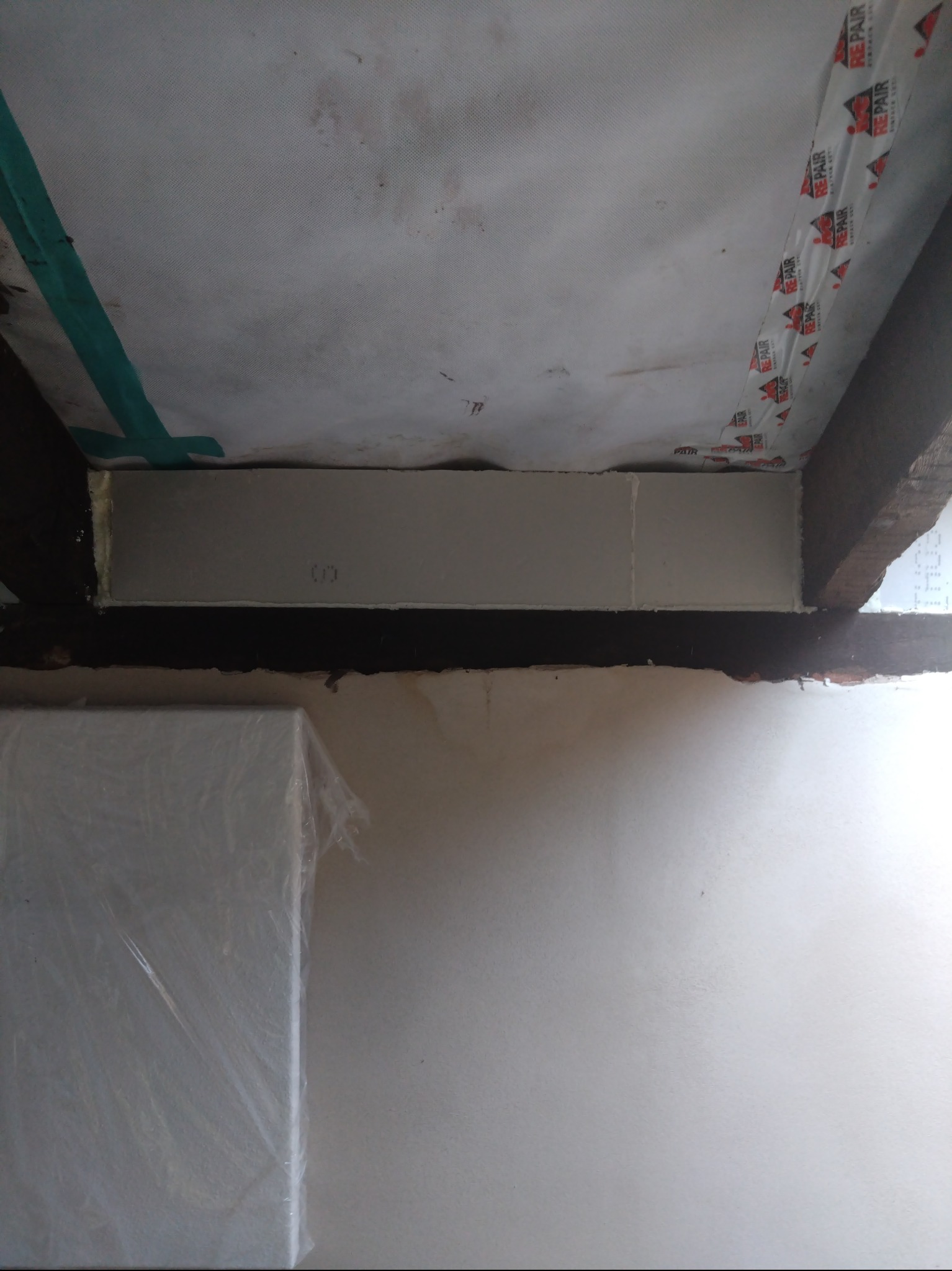 Fragment konstrukcji ściany z zamontowaną płytą gipsowo-kartonową, widoczne drewniane belki i taśma uszczelniająca.