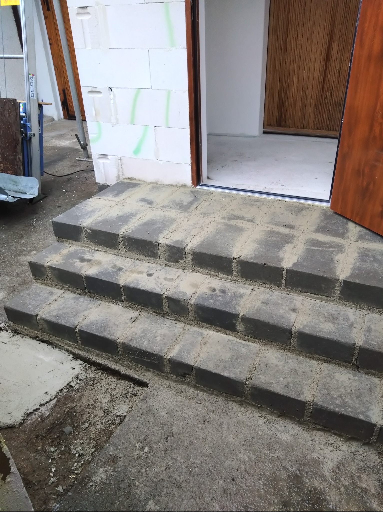 Trzy stopnie schodów wejściowych, wykonane z szarej kostki, prowadzące do drzwi z ciemnego drewna, z widocznymi śladami zaprawy i konstrukcji budynku w tle.