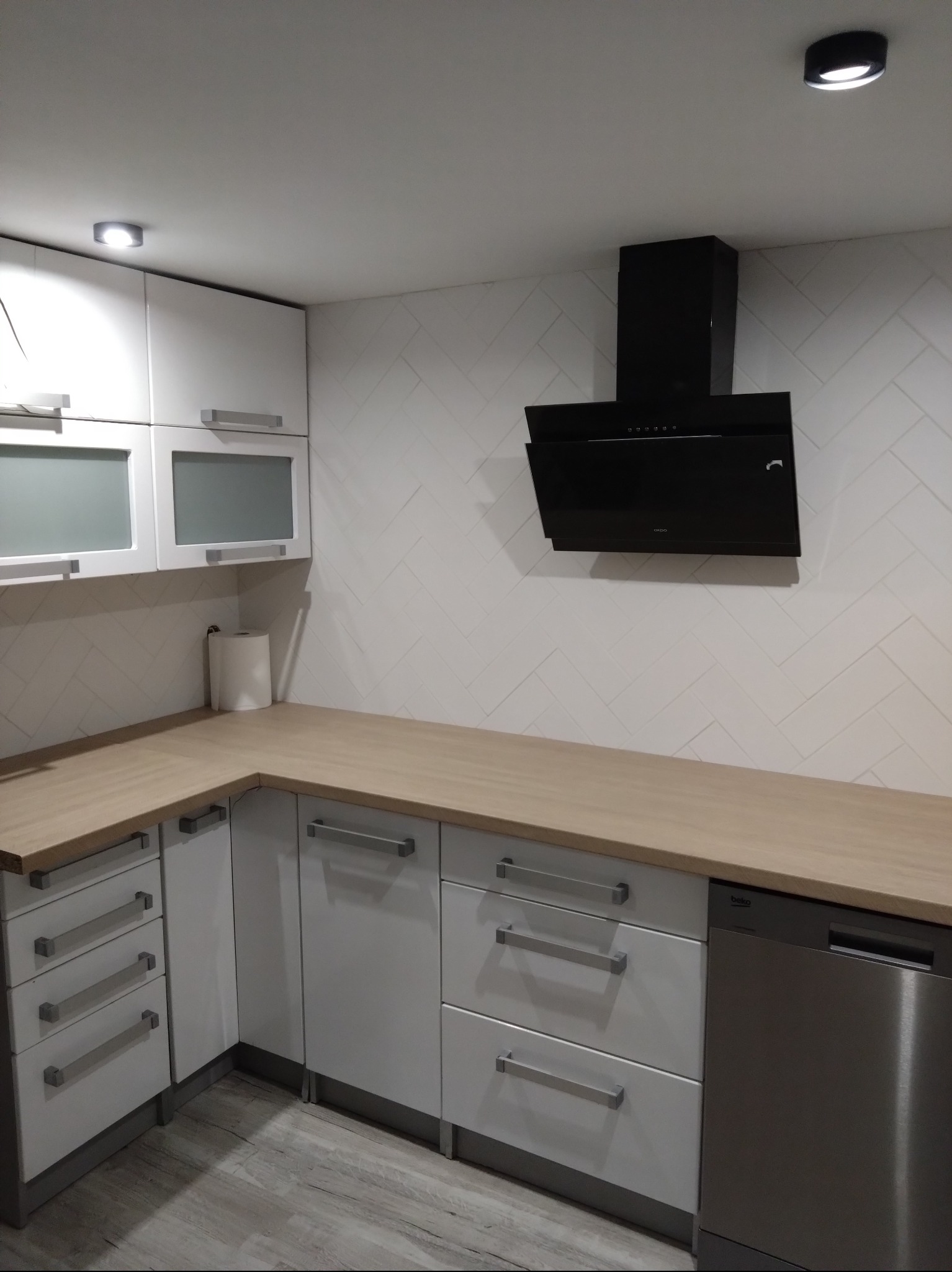 Nowoczesna kuchnia z białymi szafkami, drewnianym blatem i czarnym okapem na ścianie wyłożonej białymi płytkami w jodełkę.