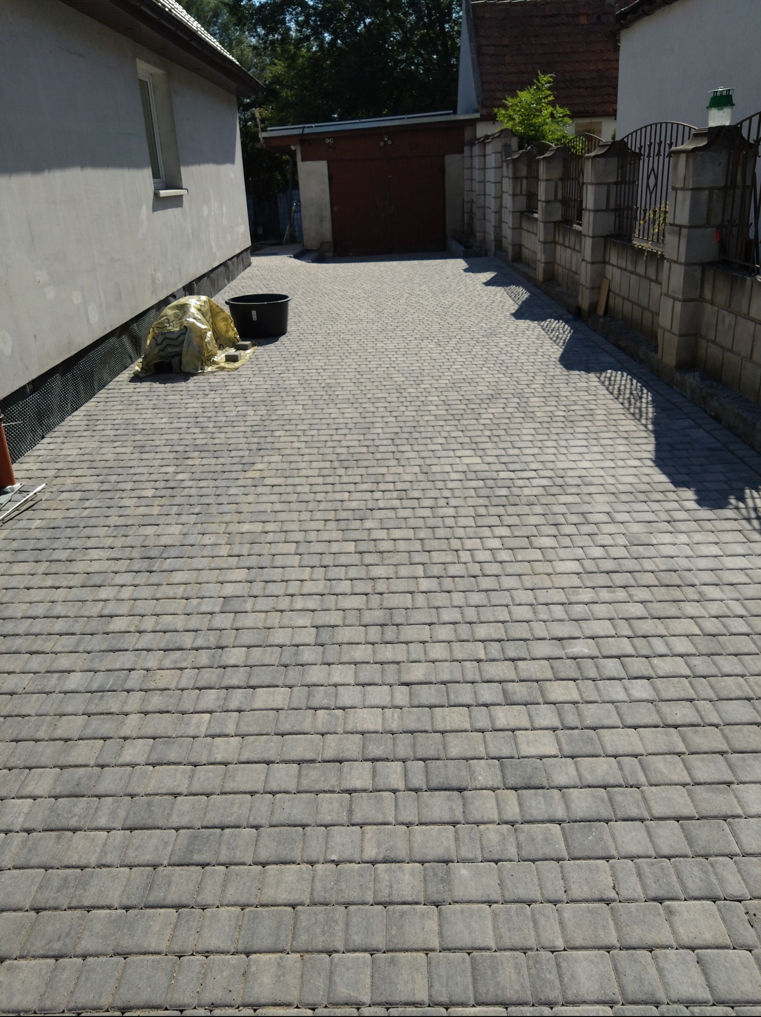 Ułożona szara kostka brukowa na podjeździe, widoczny fragment ściany budynku z ociepleniem i czarna folia ochronna, w tle garaż i fragment murowanego ogrodzenia.
