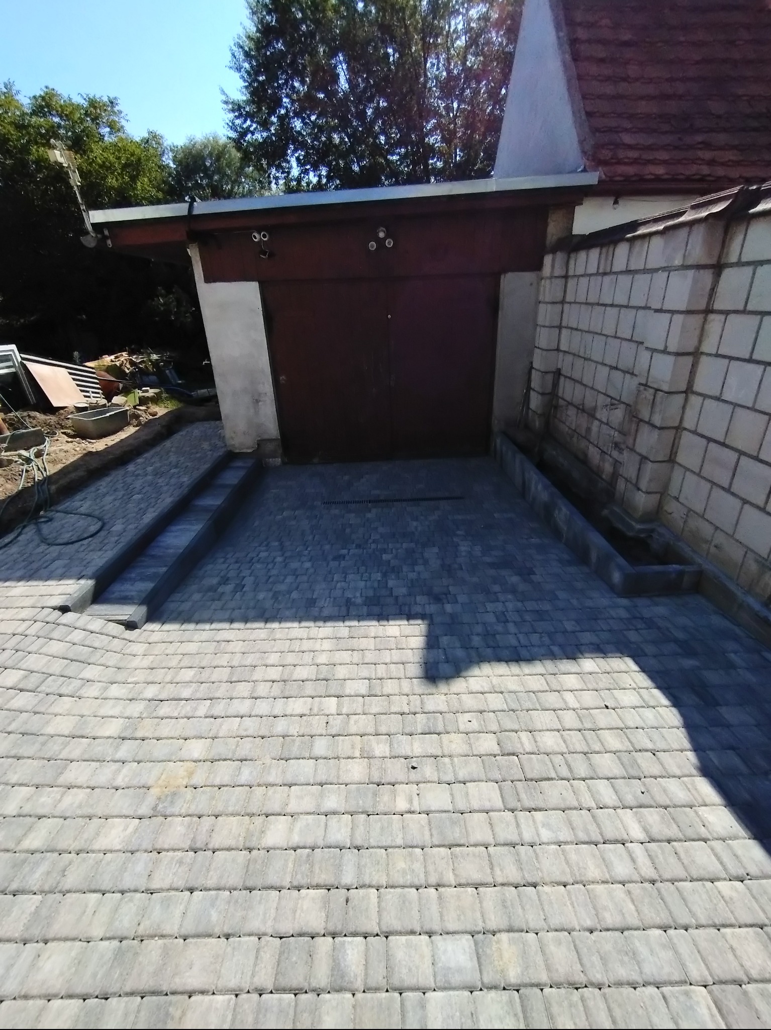 Ułożona szara kostka brukowa na podjeździe do garażu z ceglaną ścianą po prawej stronie i widocznym fragmentem dachu nad bramą garażową.