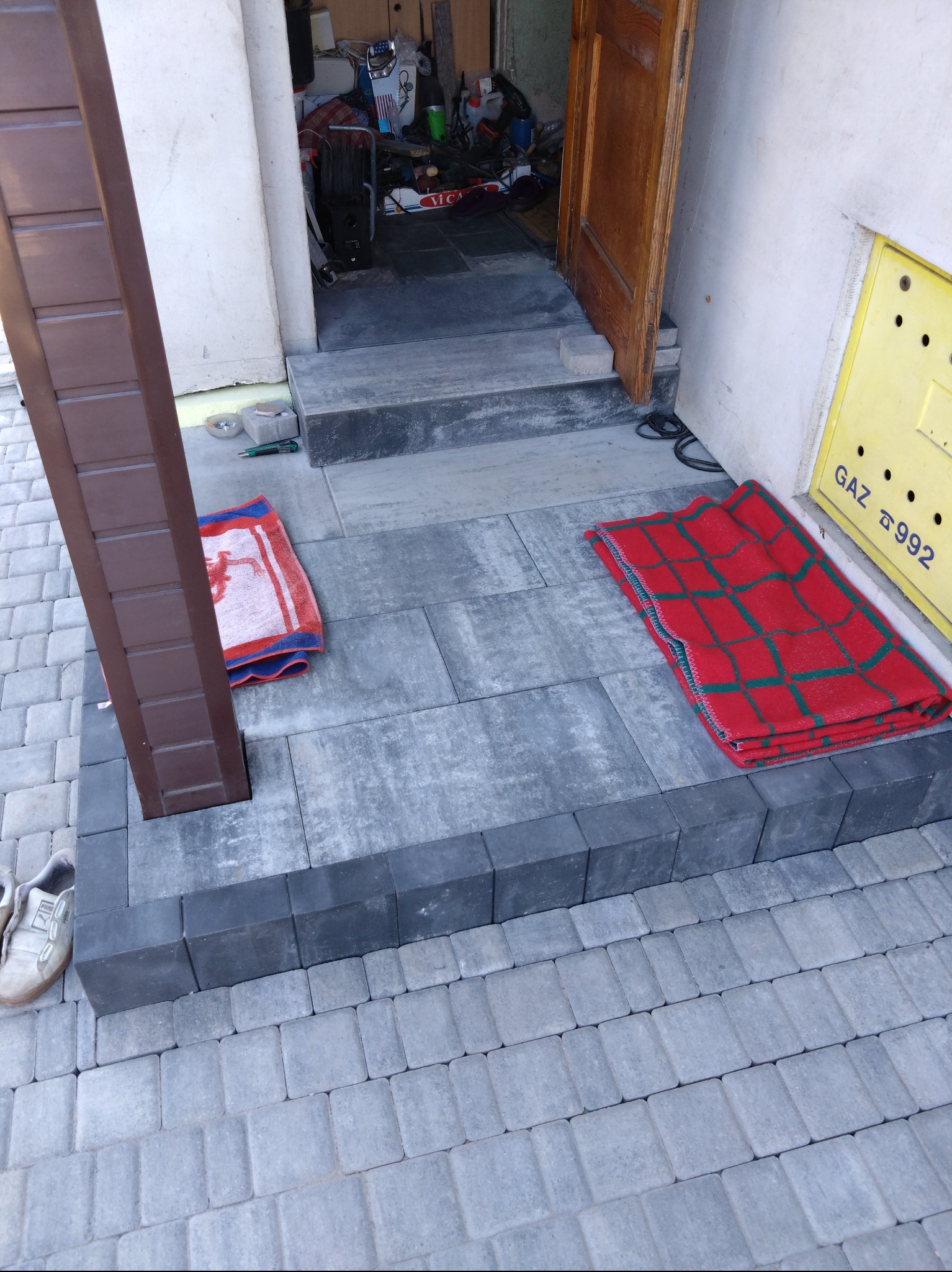 Ułożona kostka brukowa przed wejściem do budynku, z brązowym słupkiem, dywanikami i butami obok.