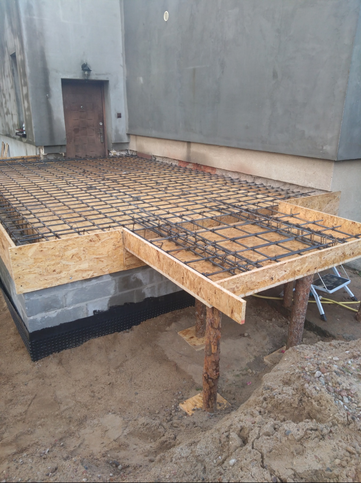 Przygotowanie zbrojenia i szalunku pod wylewkę fundamentową przy istniejącym budynku z szarą elewacją, widoczne podpory z okrąglaków.