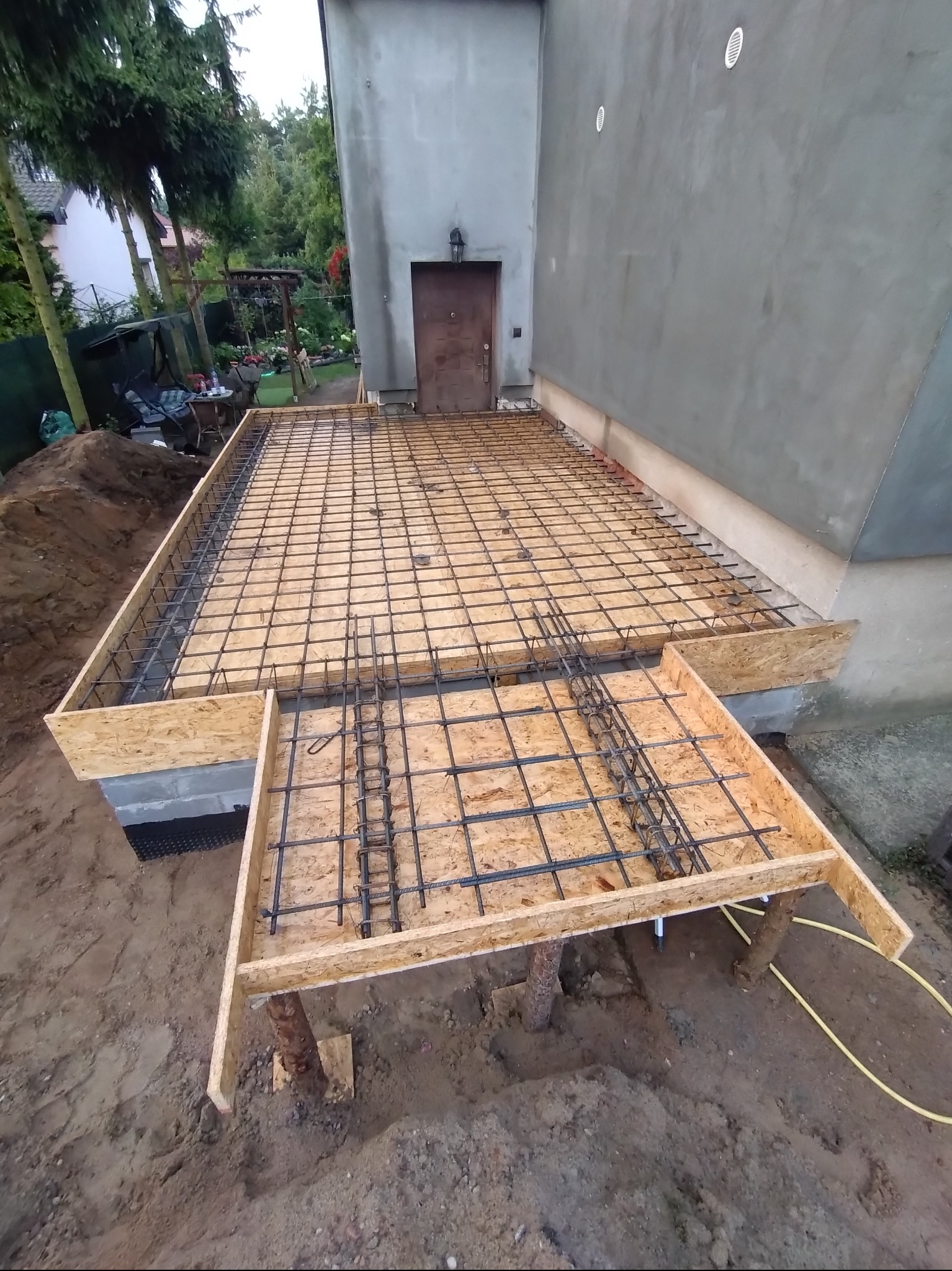 Przygotowanie szalunku i zbrojenia pod wylewkę betonową fundamentu domu jednorodzinnego, widoczne drewniane deski, stalowe pręty zbrojeniowe oraz fragment elewacji budynku.