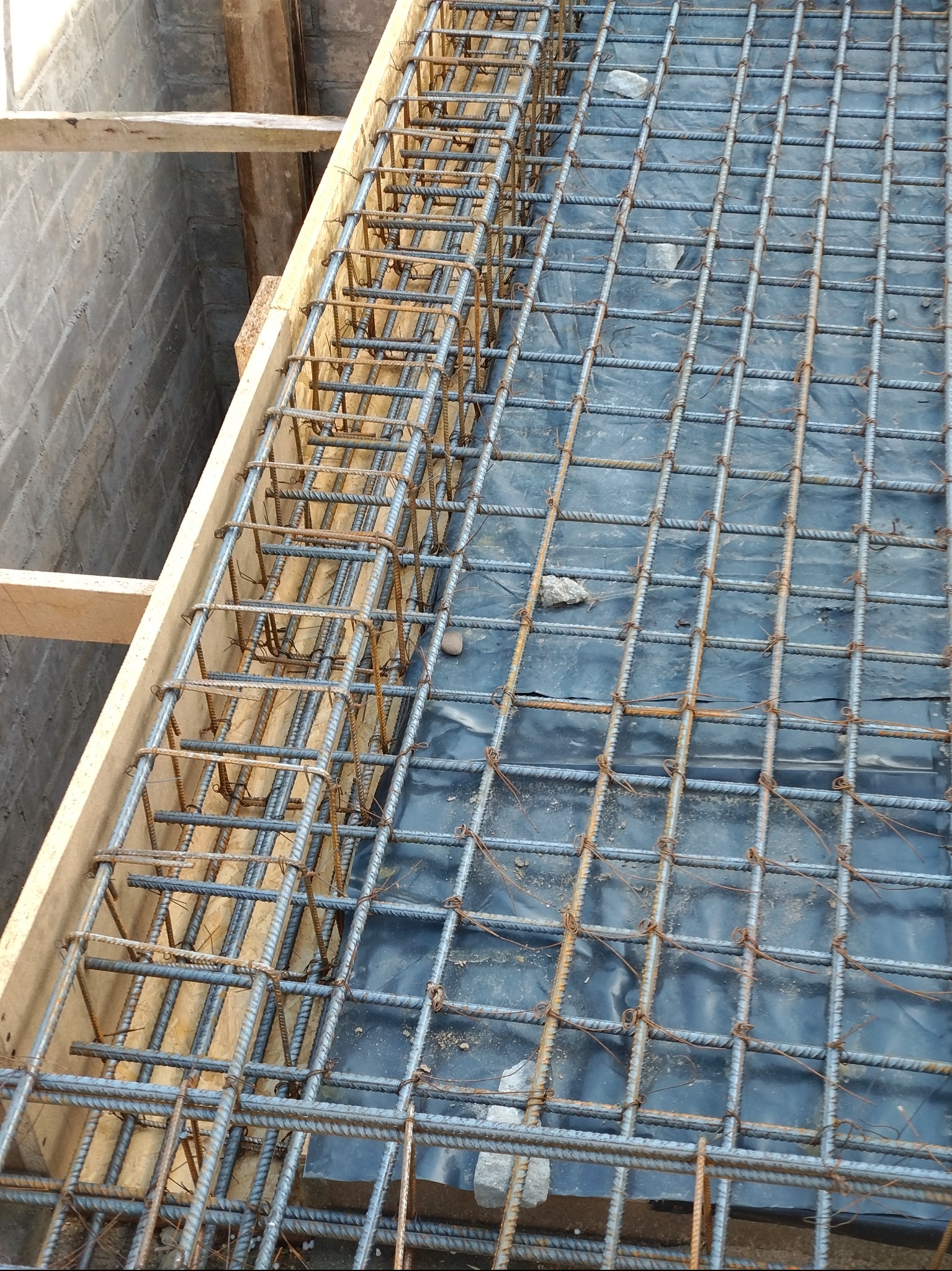 Zbrojenie fundamentów w drewnianym szalunku, widoczne stalowe pręty i czarna folia izolacyjna.