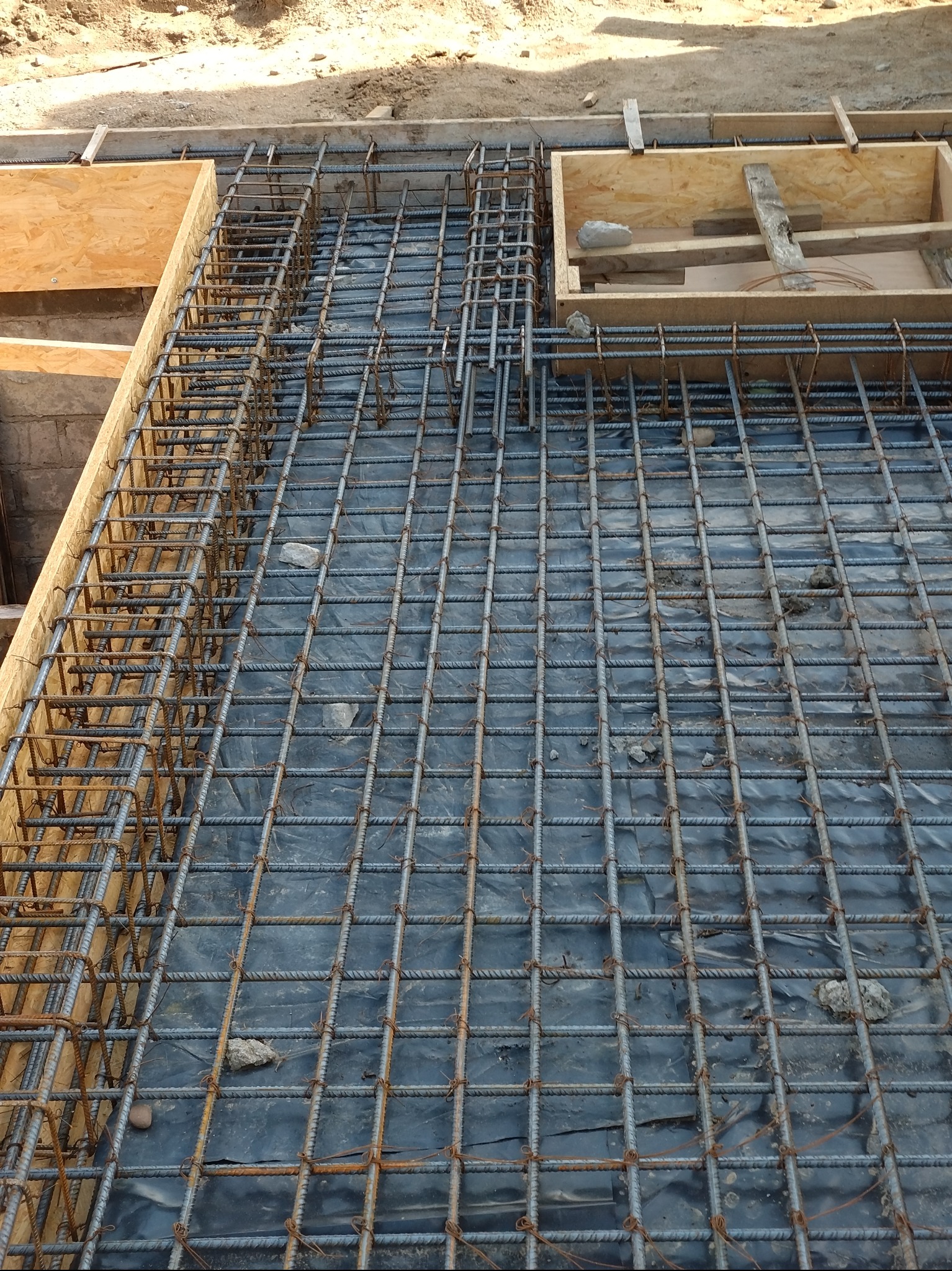Zbrojenie fundamentu z siatki stalowej, drewniany szalunek w narożnikach, czarna folia izolacyjna pod zbrojeniem, widoczne kamienie i resztki betonu.