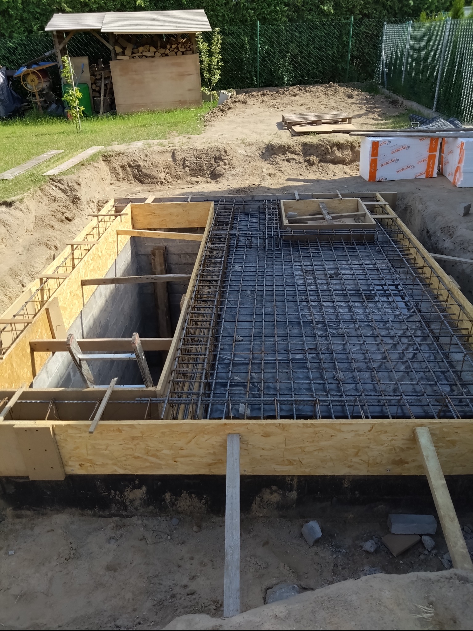 Przygotowanie zbrojenia i szalunku pod wylewkę fundamentów, widoczne wystające pręty zbrojeniowe i drewniany szalunek w wykopie.