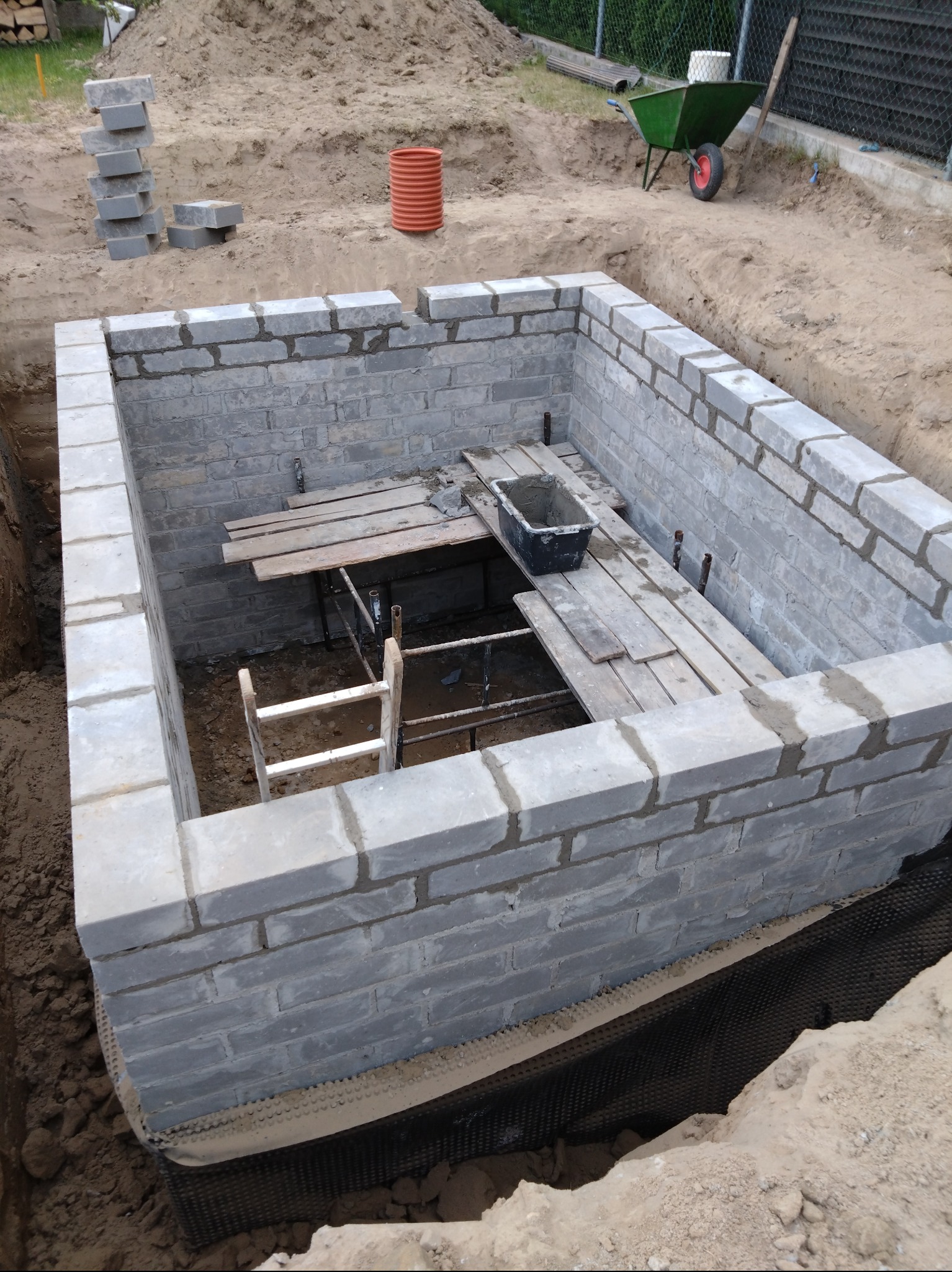 Wykop z częściowo wymurowanymi ścianami z szarych bloczków, zabezpieczony folią izolacyjną, z widocznymi deskami i drabinką w środku, oraz stertą bloczków i taczka w tle.
