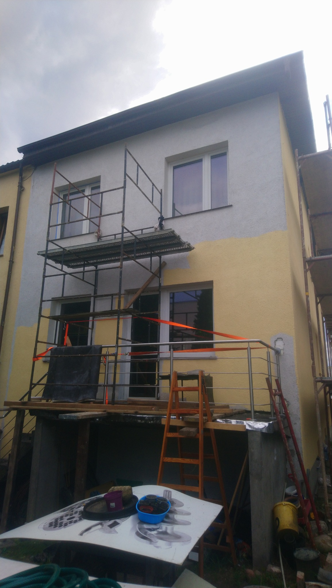 Dwupiętrowy dom w trakcie malowania elewacji na żółto, z widocznym rusztowaniem, drabinką i przygotowanymi narzędziami malarskimi.