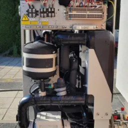 Wnętrze pompy ciepła z widocznymi przewodami, elementami sterującymi i izolacją, umieszczone na zewnątrz budynku na tle zielonych drzew i błękitnego nieba.