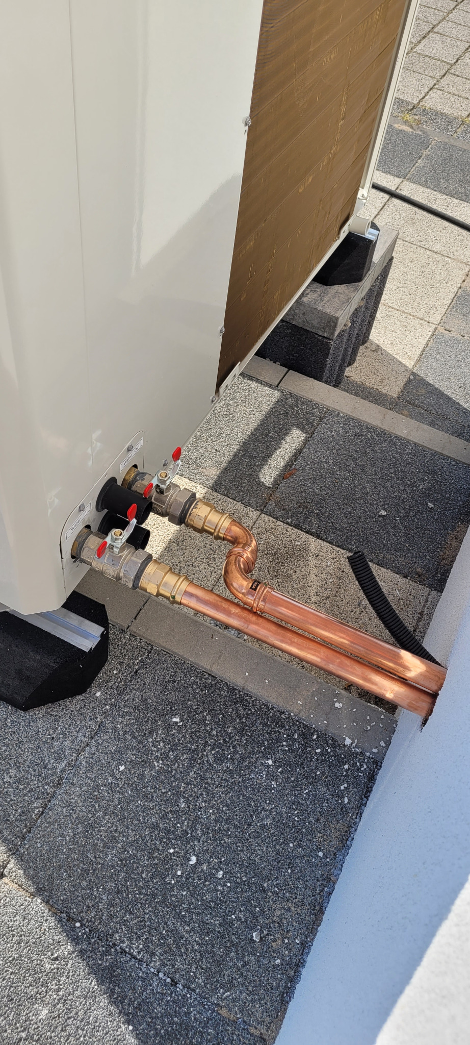 Fragment instalacji pompy ciepła z miedzianymi rurami i zaworami odcinającymi, widoczne przyłącza wody oznaczane jako 'Water In' i 'Water Out'.