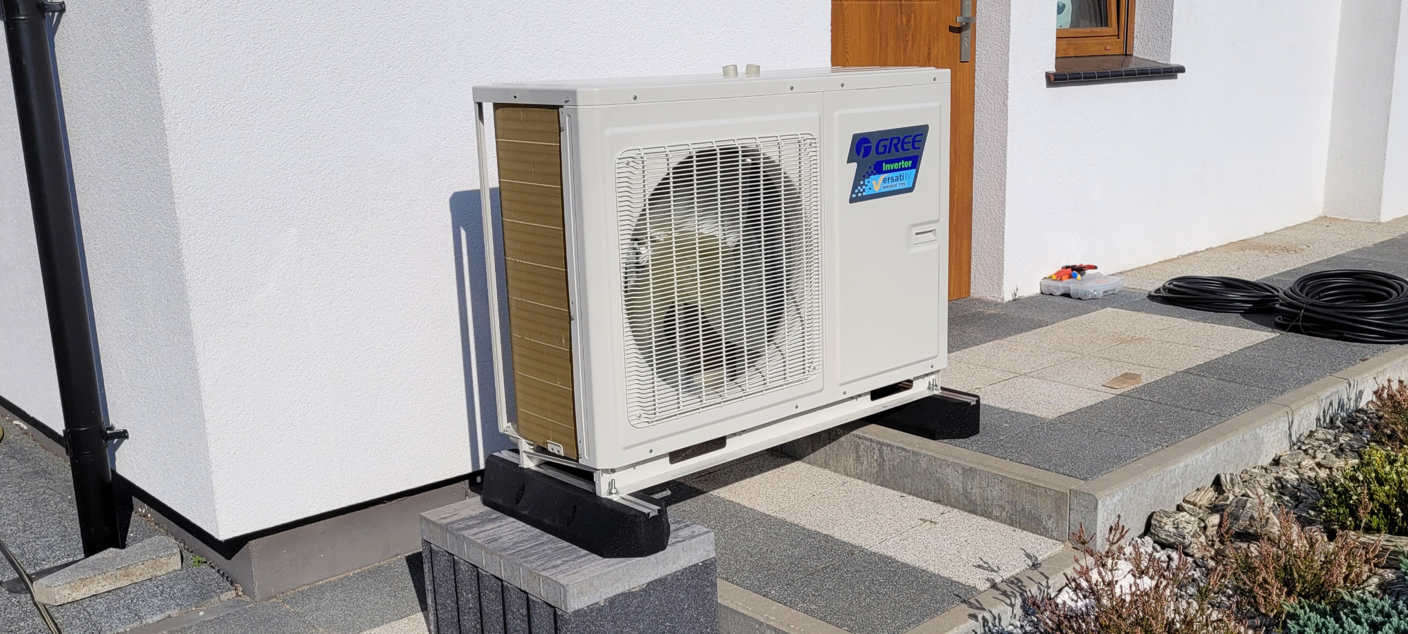 Zewnętrzna jednostka klimatyzacji marki Gree zamontowana na czarnych podstawach antywibracyjnych, na tle białej ściany budynku, obok widoczne narzędzia i zwój czarnego kabla.