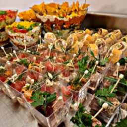 Urozmaicony bufet z przekąskami: sałatki warstwowe w szklanych misach, roladki z tortilli oraz mini porcje w przezroczystych pojemnikach, ozdobione ziołami i szpikulcami