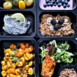 Zestaw sześciu posiłków w czarnych pojemnikach: ziemniaki z koperkiem, ryba w folii z cytryną, jogurt z jagodami, płatki zbożowe z jeżynami i lukrowanymi kawałkami, kluski gnocchi w sosie, kurczak...