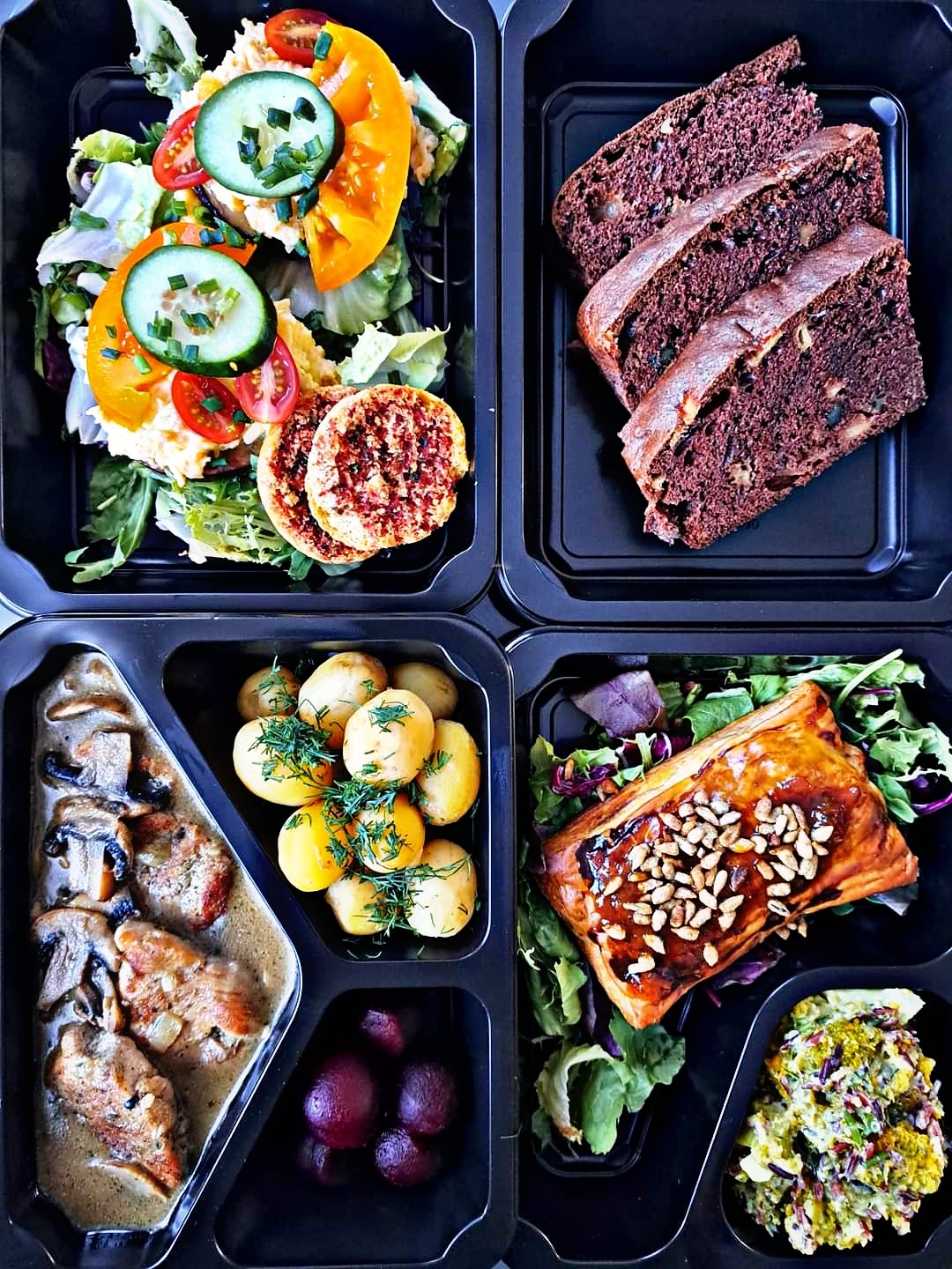 Czterodaniowy posiłek w czarnym, dzielonym pojemniku: sałatka z pomidorem i ogórkiem, ciemne ciasto z orzechami, mięso w sosie grzybowym z ziemniakami i buraczkami oraz zapiekanka z nasionami...