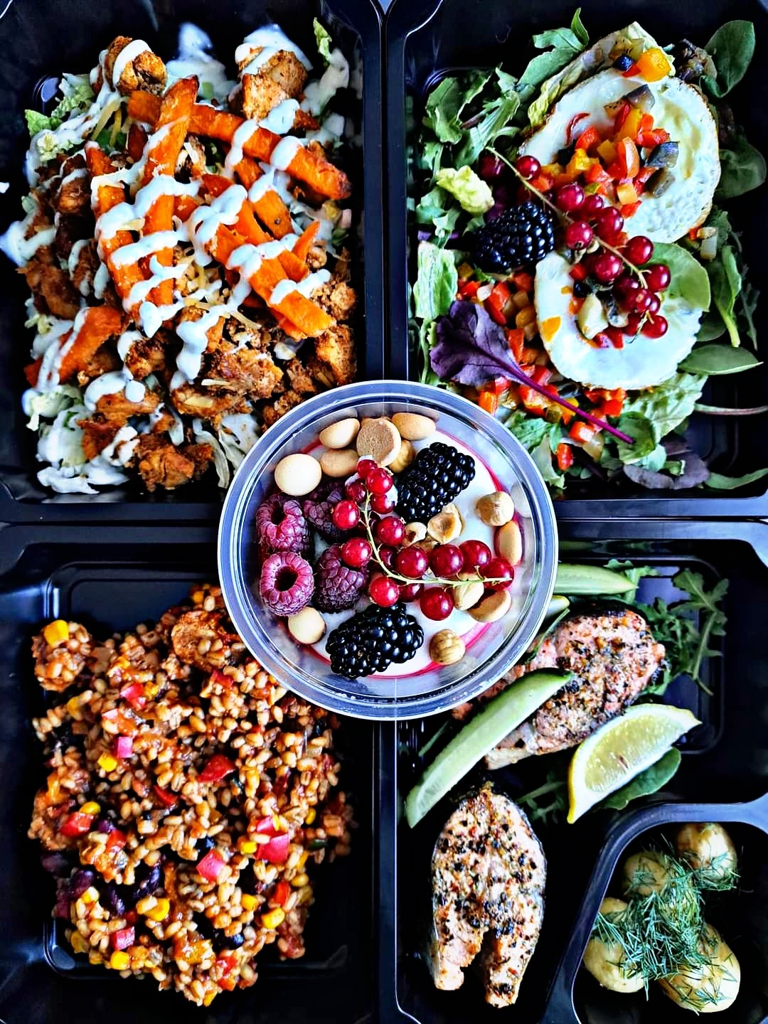 Zestaw czterech posiłków dietetycznych w czarnych pojemnikach, widok z góry: kurczak z batatami i sosem, sałatka z jajkiem i owocami leśnymi, kasza z warzywami i fasolą, łosoś z cytryną...