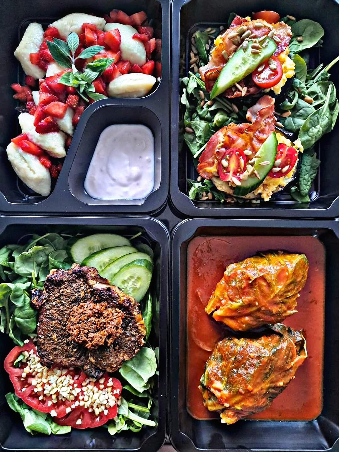 Cztery czarne pudełka z różnorodnymi daniami: pierogi z truskawkami i sosem, jajecznica z boczkiem i warzywami, stek z warzywami, gołąbki w sosie pomidorowym.