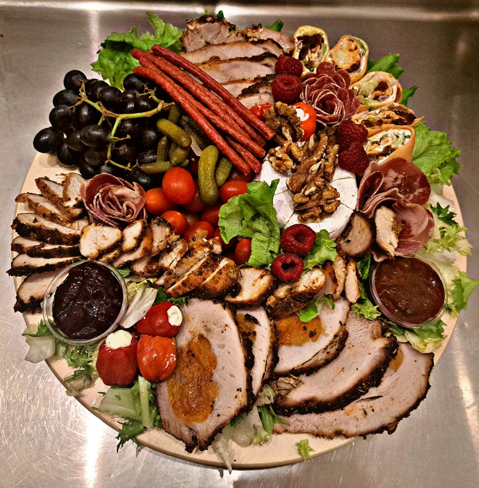 Drewniana deska z różnorodnymi przekąskami: wędliny, sery, owoce, warzywa, roladki i sosy, ułożone na sałacie.