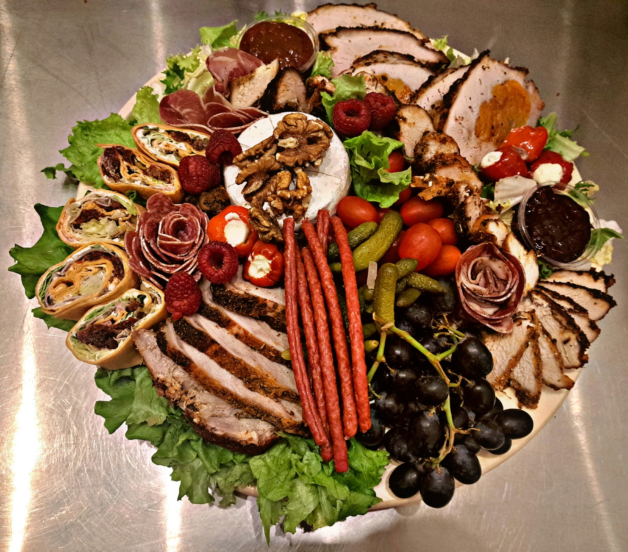 Dekoracyjna deska z przekąskami: plastry pieczonego mięsa, roladki z tortilli, salami ułożone w kształt róży, ser brie z orzechami, winogrona, ogórki konserwowe, pomidorki koktajlowe, papryczki...