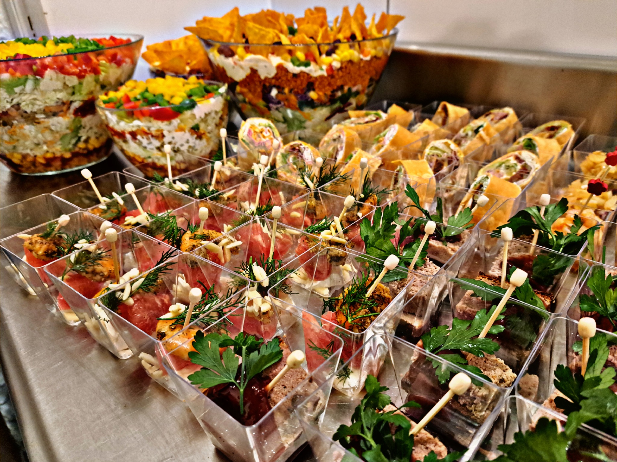 Urozmaicony bufet z przekąskami: sałatki warstwowe w szklanych misach, roladki z tortilli oraz mini porcje w przezroczystych pojemnikach, ozdobione ziołami i szpikulcami
