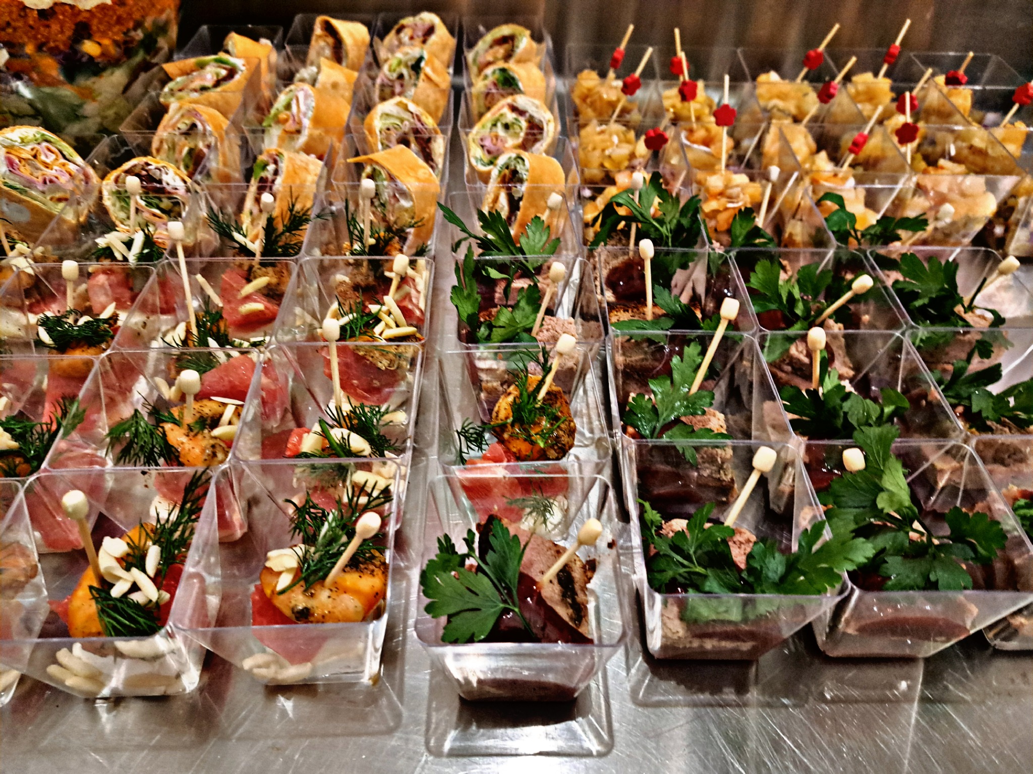 Różnorodne przekąski cateringowe w przezroczystych, kwadratowych pojemnikach: roladki z tortilli, koreczki z mięsem i warzywami, ozdobione ziołami i drewnianymi wykałaczkami.
