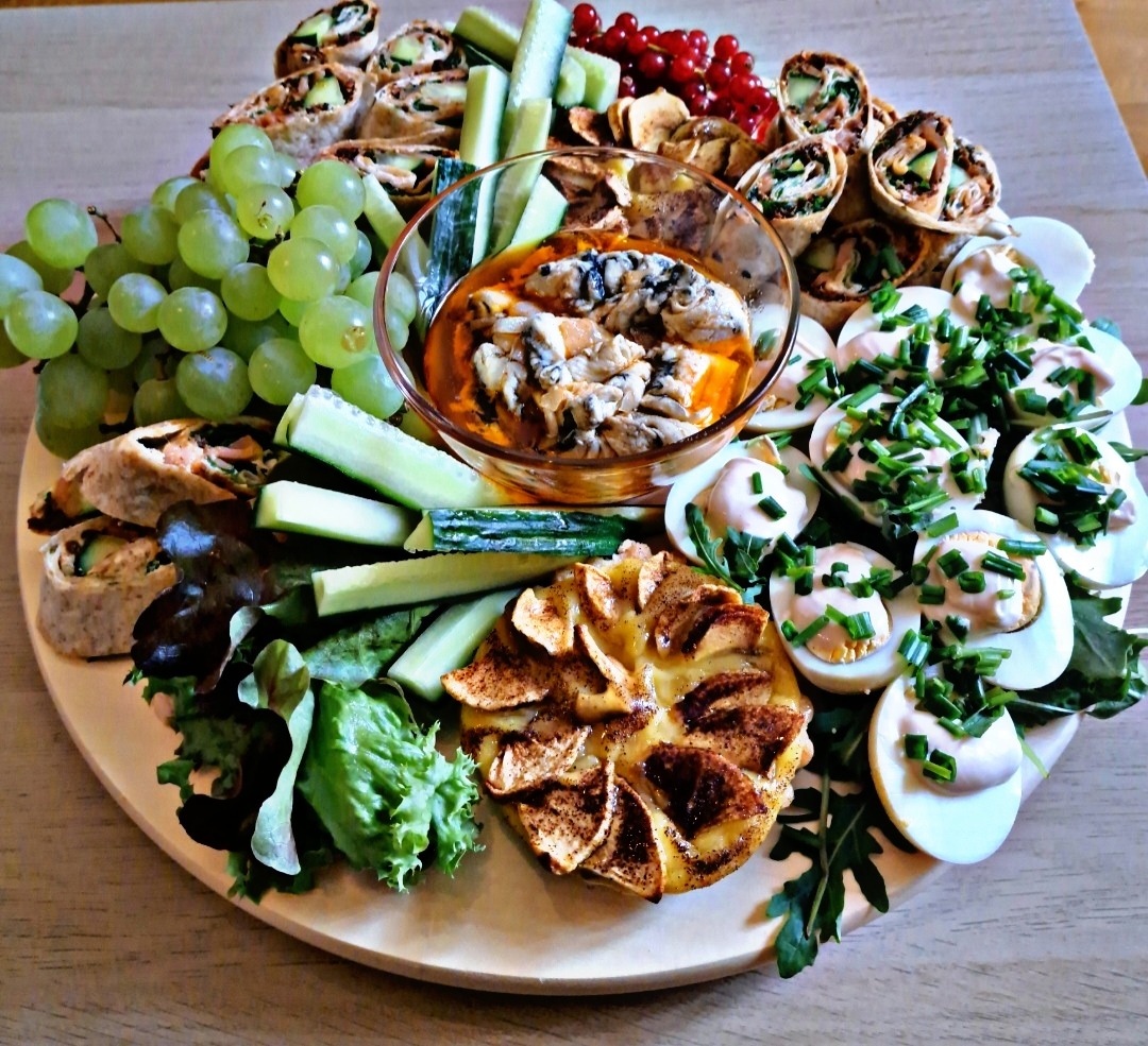 Dekoracyjna drewniana taca z przekąskami: roladki tortilli z warzywami, plastry ogórka, zielone winogrona, czerwone porzeczki, jajka na twardo posypane szczypiorkiem, sałata, tarta jabłkowa...