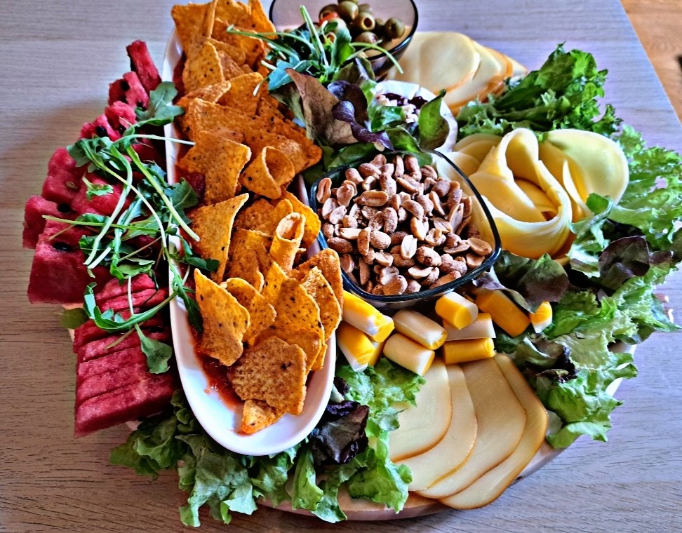 Deska przekąsek z arbuzem, nachos z sosem, orzechami, serem, paluszkami krabowymi i oliwkami, udekorowana sałatą i rukolą.