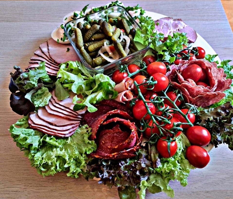 Dekoracyjna taca z wędlinami, pomidorkami koktajlowymi, sałatą i korniszonami w szklanym naczyniu, ułożona na drewnianym stole.