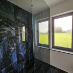 Nowoczesna łazienka z kabiną prysznicową typu walk-in wykończona czarnymi płytkami imitującymi marmur z niebieskimi żyłami, dwa okna z widokiem na zielone pole i panele słoneczne.
