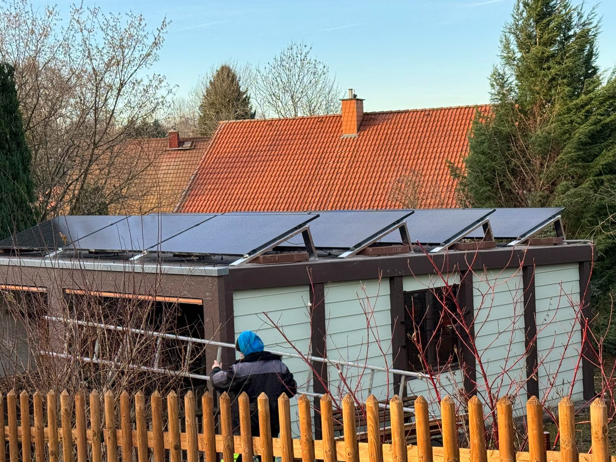 Instalacja paneli słonecznych na dachu garażu, widok zza drewnianego płotu, pracownik na drabinie w niebieskiej czapce.
