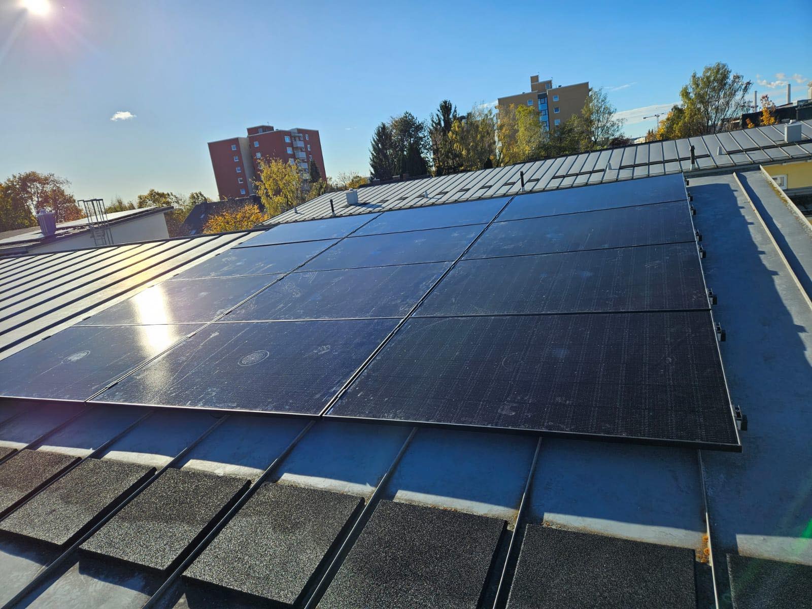 Panele słoneczne zamontowane na metalowym dachu budynku, widoczne budynki mieszkalne w tle pod błękitnym niebem.