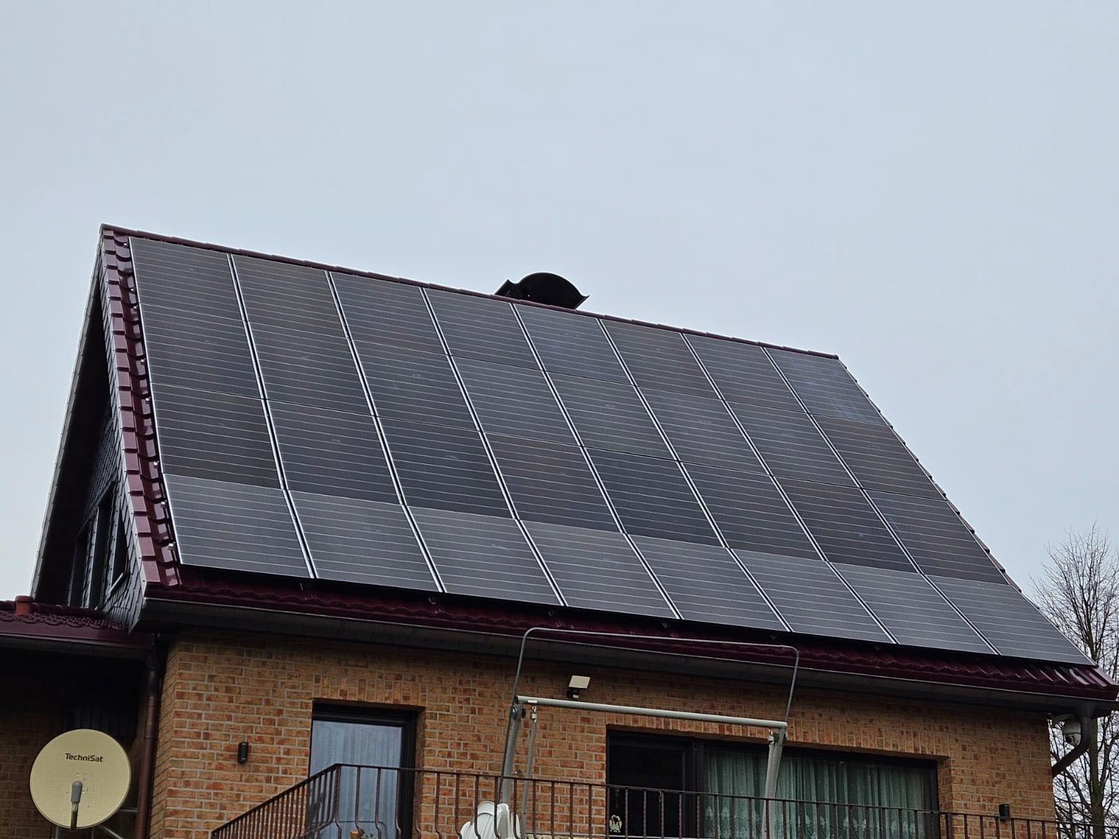 Panele słoneczne na skośnym dachu ceglanego domu jednorodzinnego, widoczne okna, balkon i antena satelitarna TechniSat.