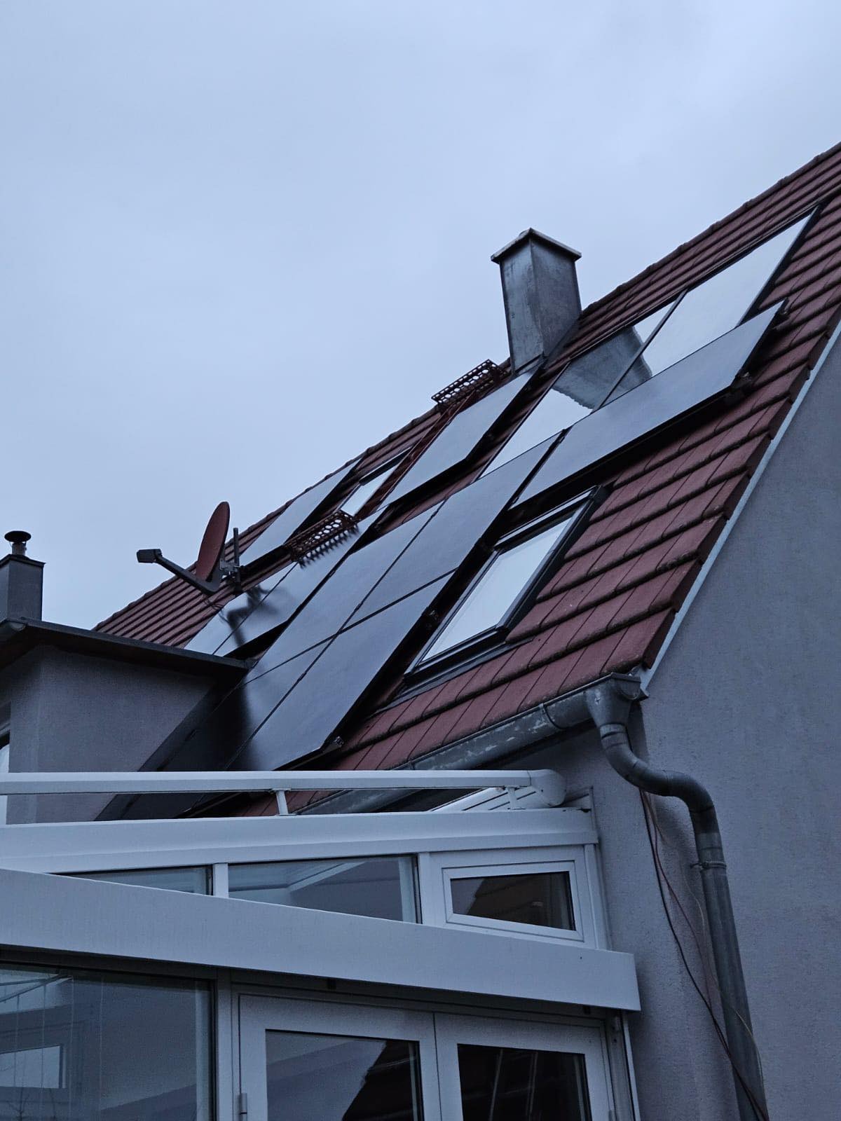 Czerwony dach z panelami słonecznymi, kominem i anteną satelitarną, widziany pod kątem z dołu, w pochmurny dzień.