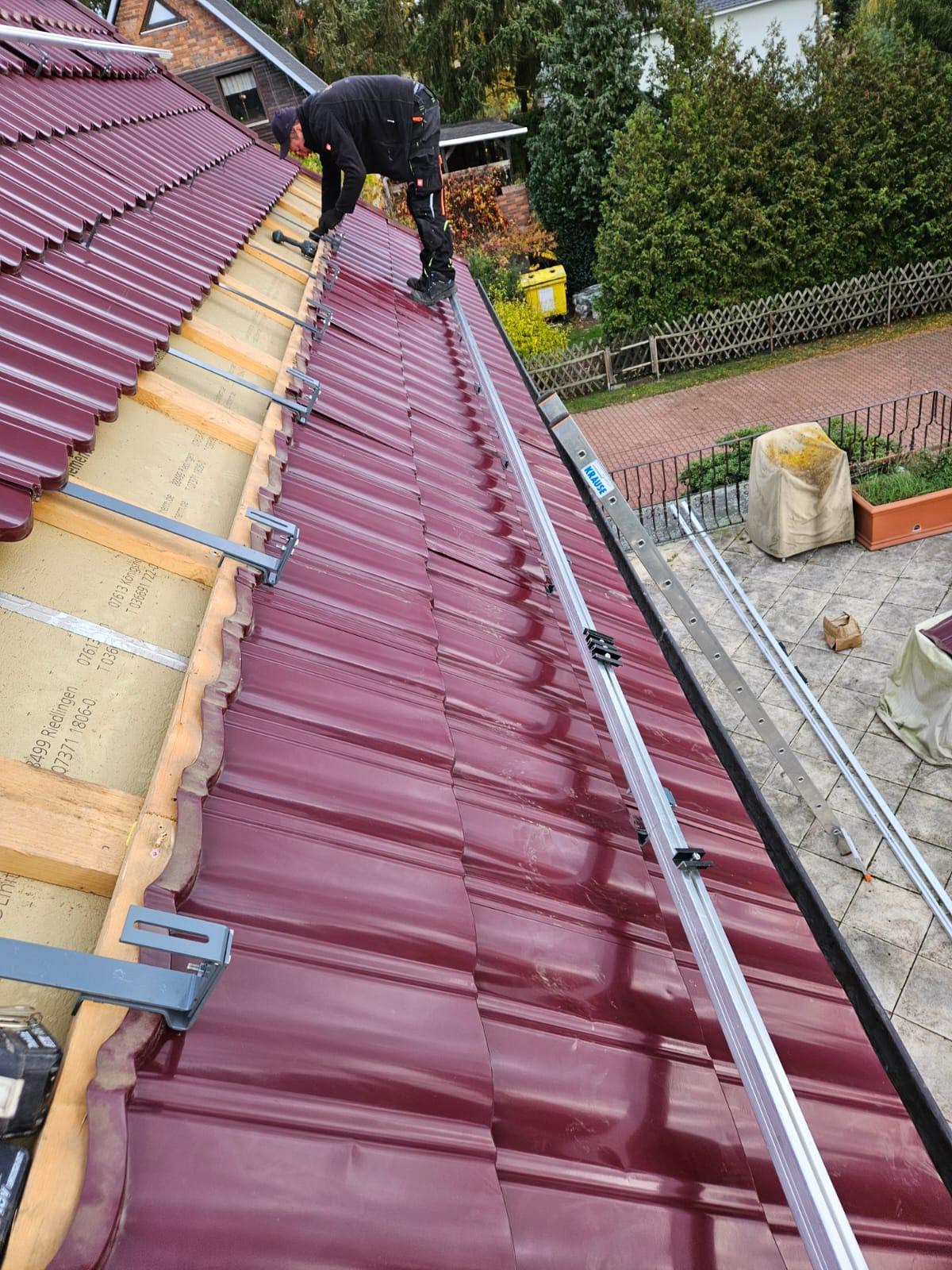 Montaż konstrukcji wsporczej pod panele słoneczne na dachu pokrytym bordową dachówką, widoczne aluminiowe profile i uchwyty mocujące, pracownik w kombinezonie na dachu, drabina Krause z boku.