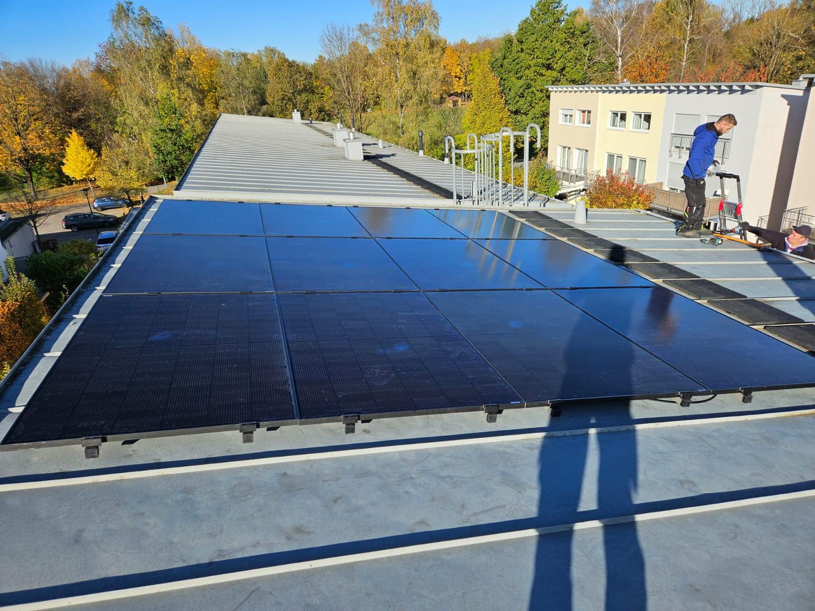 Montaż paneli słonecznych na płaskim dachu budynku, widoczne narzędzia i dwóch instalatorów, jesienne drzewa w tle.
