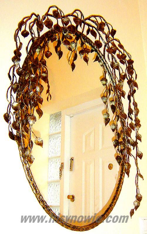 Owalne lustro z kutą, metalową ramą w kształcie gałęzi z liśćmi, odbijające białe drzwi z szybą z mlecznego szkła.