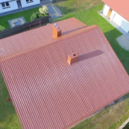 Widok z góry na budynek z nowym, brązowym dachem z blachy trapezowej i dwoma kominami, otoczony zielonym trawnikiem i innymi budynkami.