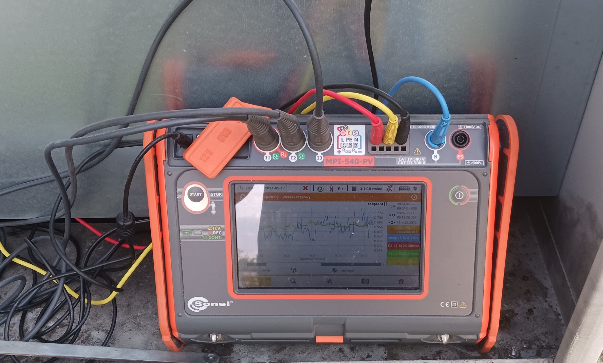 Analizator jakości zasilania Sonel MPI-540-PV z podłączonymi przewodami pomiarowymi, wyświetlający wykres czasowy z danymi pomiarowymi, umieszczony w szarej, metalowej obudowie. Widoczna naklejka...