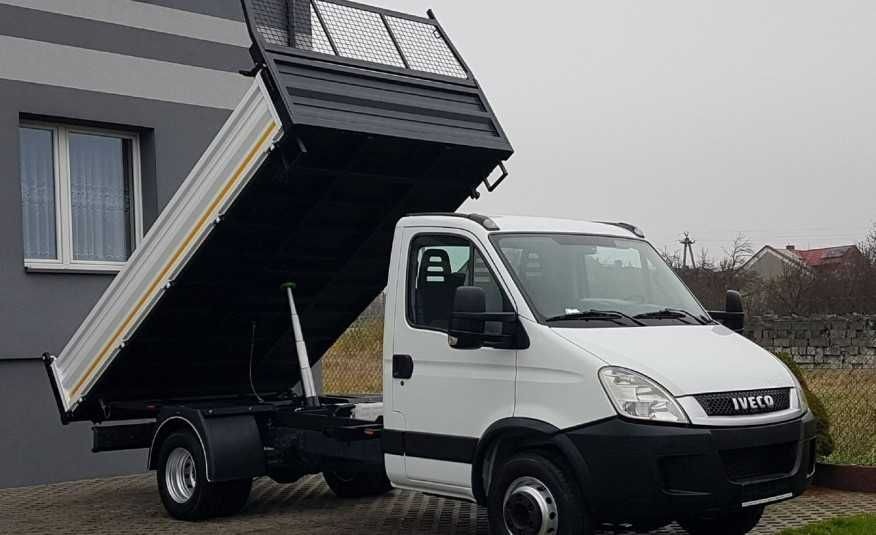 Biały samochód ciężarowy marki Iveco z podniesioną skrzynią ładunkową, widoczny na tle szarego budynku i zarośli.