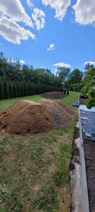 Trzy pryzmy materiałów budowlanych: piasek, żwir i ziemia, ułożone na trawniku w ogrodzie, na tle żywopłotu z tuj i błękitnego nieba z obłokami.