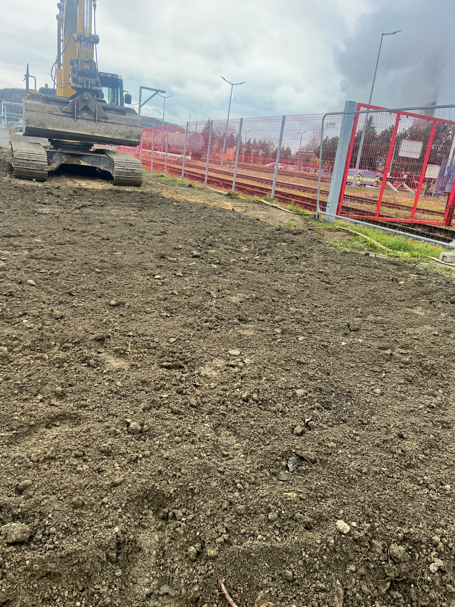 Żółta koparka gąsienicowa przygotowuje teren pod budowę, z widocznymi torami kolejowymi w tle za czerwonym ogrodzeniem.