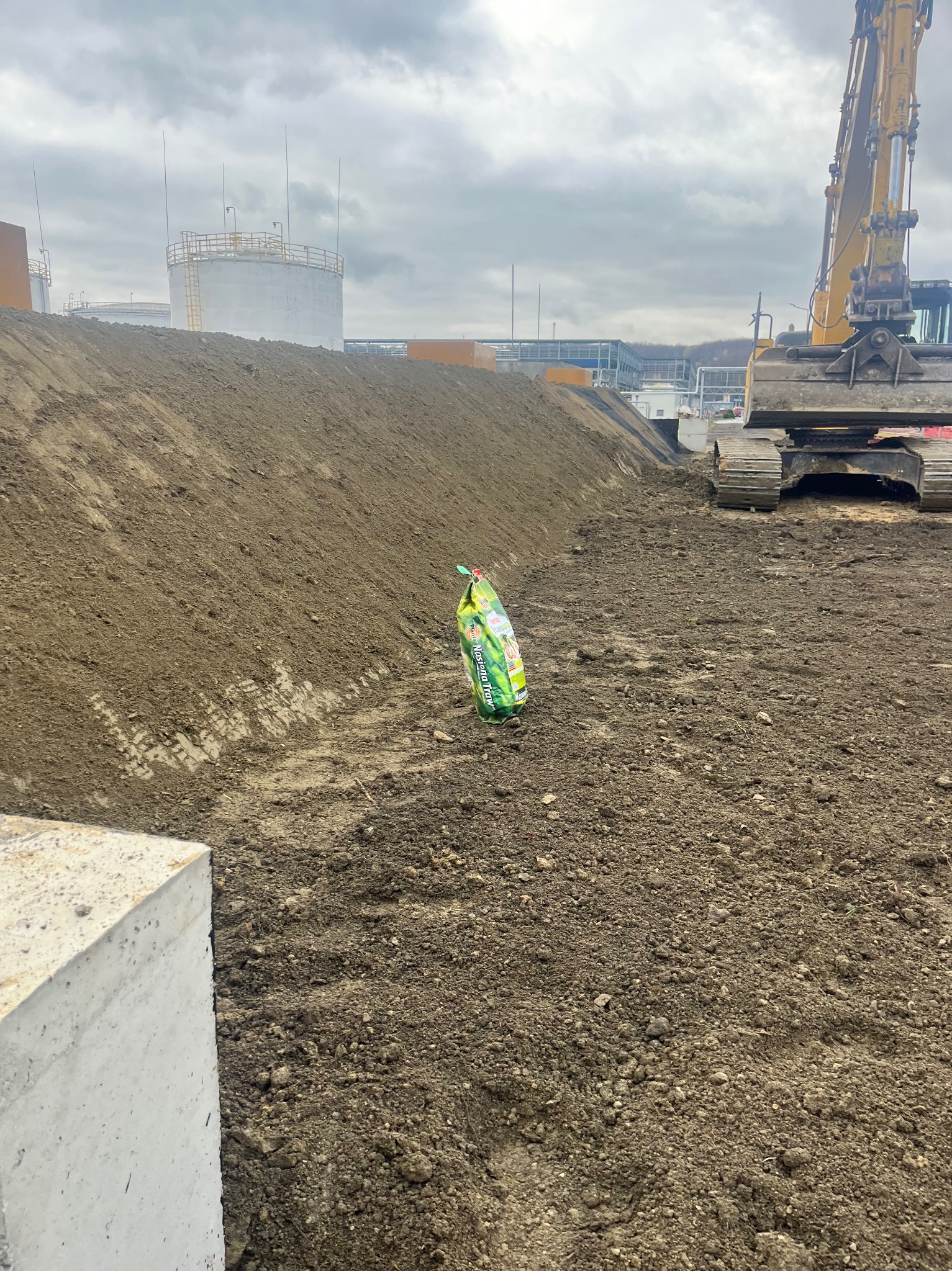 Żółta koparka w trakcie prac ziemnych na placu budowy, obok leży worek z ziemią, w tle widoczne zbiorniki przemysłowe i pochmurne niebo.