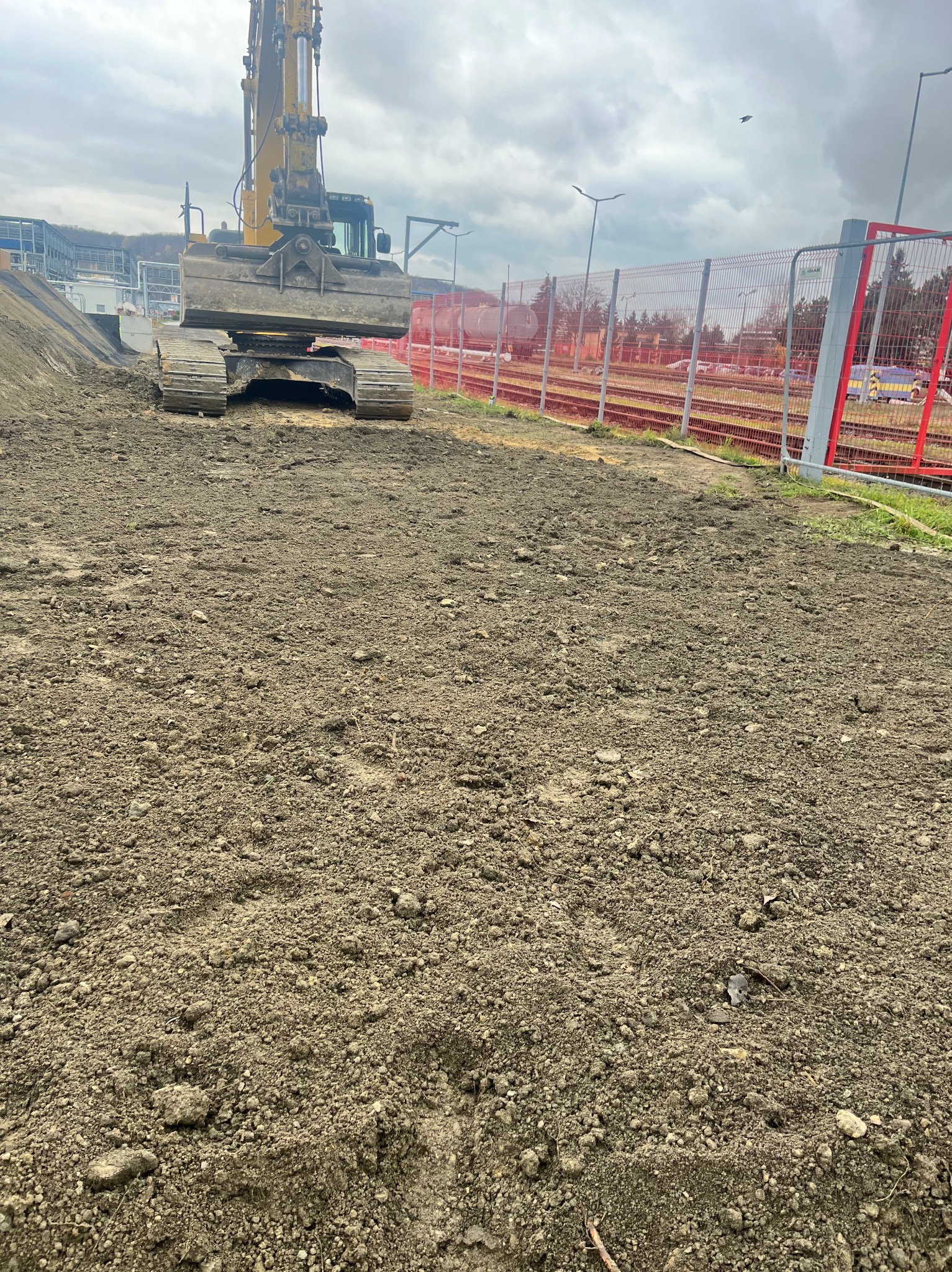 Żółta koparka gąsienicowa na placu budowy, widok z perspektywy gruntu, w tle ogrodzenie z siatki i zabudowania kolejowe.