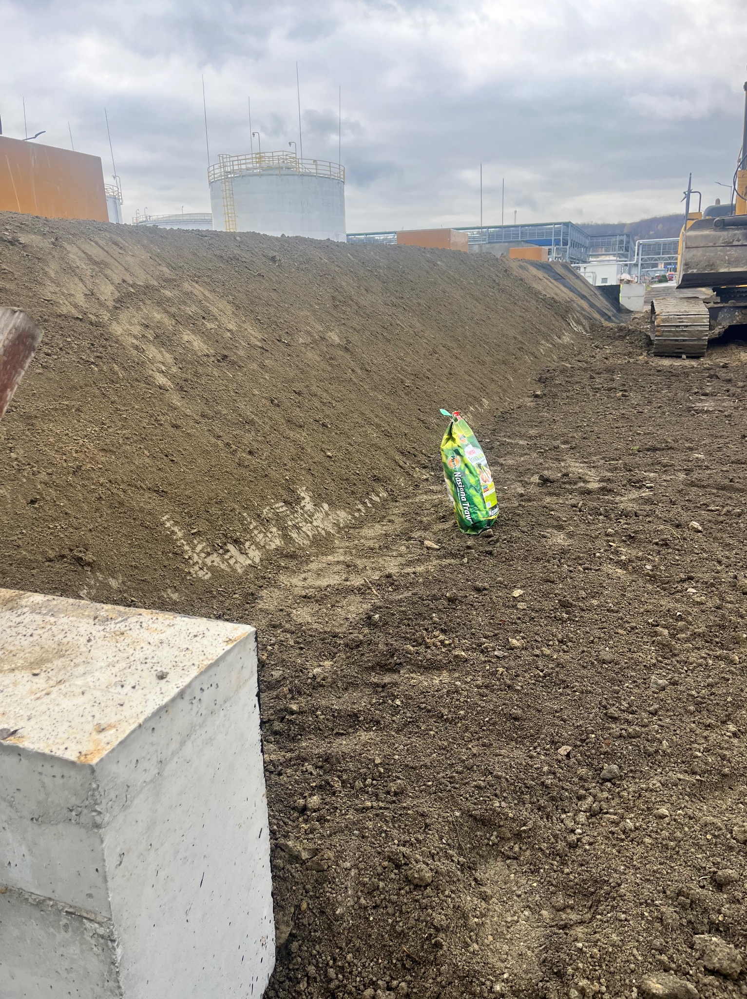 Przygotowany teren pod budowę z widocznym wykopem, stertą ziemi, workiem nawozu i fragmentem koparki w tle, zbiorniki przemysłowe w oddali.