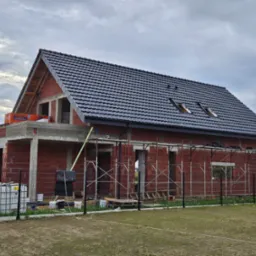 Surowy budynek w trakcie budowy z częściowo ułożonym, ciemnym dachem z dach&oacute;wki, widoczne rusztowania i materiały budowlane oraz pracownik na dachu.