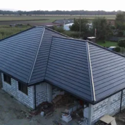 Nowy, grafitowy dach z blachodachówki modułowej na surowym budynku z białego pustaka, widok z góry na tle pól i zabudowań.