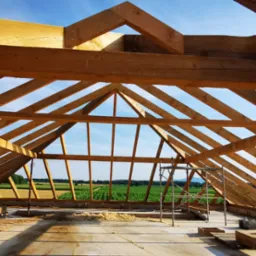 Konstrukcja drewniana dachu w trakcie montażu, widok na otwartą przestrzeń i pola uprawne przez szkielet dachu, ze stertą drewna na pierwszym planie i rusztowaniem w środku kadru.